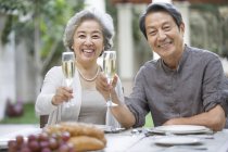 Senior pareja china sosteniendo flautas de champán en la mesa de comedor - foto de stock
