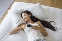 Chinese woman lying on bed with cup of coffee — Stock Photo