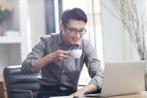Азиатский человек, работающий с ноутбуком и кофе в офисе — стоковое фото