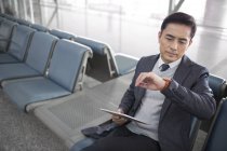 Hombre asiático esperando en el aeropuerto con tableta digital y mirando el reloj - foto de stock