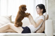 Jeune femme chinoise jouer avec animal caniche sur canapé — Photo de stock