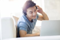 Chinese man in headphones using laptop on sofa — Stock Photo