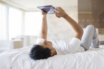 Uomo cinese sdraiato con tablet digitale sul letto — Foto stock