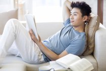 Jovem chinês homem ler livro no sofá — Fotografia de Stock