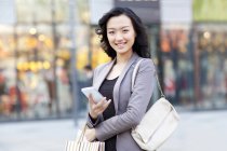 Chinese woman standing on street with shopping bag and smartphone — Stock Photo