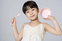 Маленькая китаянка держит миску и ложку на сером фоне — стоковое фото