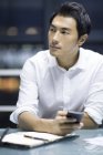Hombre de negocios chino sosteniendo el teléfono inteligente en la oficina y mirando hacia otro lado - foto de stock