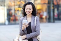 Mujer china comprobando teléfono inteligente en la calle con bolsa de compras - foto de stock