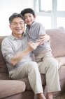 Avô chinês e neto abraçando no sofá e sorrindo — Fotografia de Stock