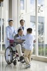 Petit-fils chinois courant vers grand-père en fauteuil roulant avec des médecins — Photo de stock