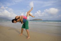 Joven hombre chino levantando mujer espalda con espalda en la playa de Hainan - foto de stock
