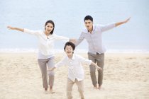 Pais chineses com filho correndo com os braços estendidos na areia da praia — Fotografia de Stock