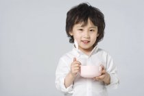Little Asian boy holding bowl and spoon on gray background — Stock Photo