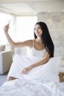 Китайська жінка, що прийняття selfie з смартфон на ліжку — стокове фото