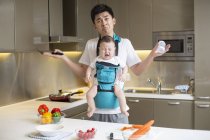 Pai chinês segurando bebê chorando e encolhendo os ombros na cozinha — Fotografia de Stock