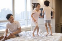 Chinesische Geschwister springen und haben Spaß auf dem Bett mit Vater — Stockfoto