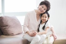 Chinese grandmother and granddaughter hugging on sofa and looking in camera — Stock Photo