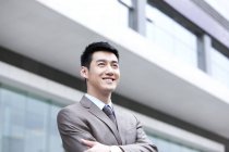 Empresário chinês sorridente com armas cruzadas em frente ao prédio de negócios — Fotografia de Stock