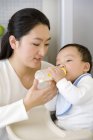 Chinesin füttert Baby-Sohn mit Flasche im Hochstuhl in Küche — Stockfoto