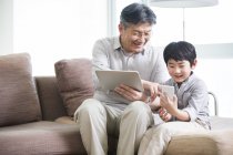 Chinese grandfather and grandson using digital tablet and smartphone on couch — Stock Photo