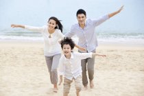 Padres chinos con hijo corriendo con los brazos extendidos en la arena de la playa - foto de stock