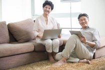 Pareja de ancianos chinos con ordenador portátil y tableta digital en la sala de estar - foto de stock