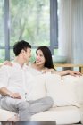 Jovem casal chinês sorrindo e olhando um para o outro no sofá — Fotografia de Stock