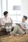 Pareja de ancianos chinos con portátil y tableta digital hablando en la sala de estar - foto de stock