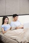 Jovem casal chinês usando dispositivos sem fio na cama — Fotografia de Stock