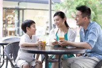 Padres chinos con hijo disfrutando de bebidas frías en la cafetería de la acera - foto de stock