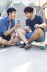 Chinese men sitting on skateboards and looking at smartphone — Stock Photo
