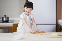 Chica china rodando masa en la mesa de la cocina y mirando en la cámara - foto de stock