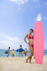 Mulher chinesa de pé com prancha de surf e amigos no fundo — Fotografia de Stock