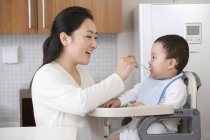 Chinês mulher alimentando bebê filho em cadeira alta na cozinha — Fotografia de Stock