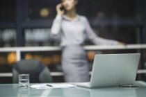 Ноутбук на рабочем столе с деловой женщиной, разговаривающей по телефону в фоновом режиме — стоковое фото