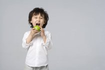 Pequeño chico asiático mordiendo manzana verde sobre fondo gris - foto de stock