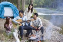Amigos chinos sentados alrededor de fogata y tocando la guitarra - foto de stock