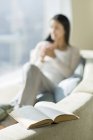 Close-up of open book with woman sitting on sofa in background — Stock Photo