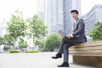 Homem de negócios chinês sentado com laptop no banco de rua — Fotografia de Stock