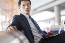 Hombre de negocios chino sentado con portátil en la sala de espera del aeropuerto - foto de stock