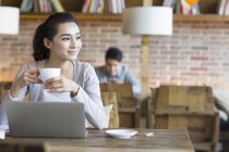 Китаянка сидит с ноутбуком и кофе в кафе — стоковое фото