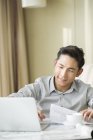 Homem chinês trabalhando em casa com papéis e laptop — Fotografia de Stock