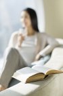Nahaufnahme eines aufgeschlagenen Buches mit einer Frau, die auf dem Sofa im Hintergrund sitzt — Stockfoto