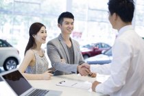 Pareja china estrechando la mano con concesionario de coches en sala de exposición - foto de stock