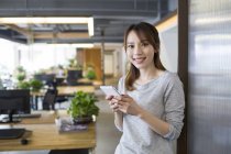 Chinese woman holding smartphone and smiling in office — Stock Photo