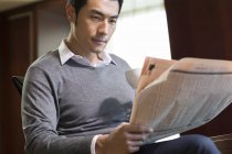 Chinese man with coffee cup reading newspaper in home interior — Stock Photo