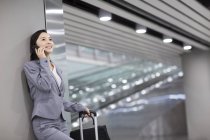 Chinese businesswoman leaning on wall and talking on phone in airport — Stock Photo
