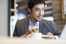 Chinesischer Geschäftsmann sitzt mit Croissant und schaut auf Laptop — Stockfoto