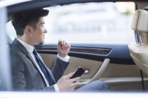 Hombre de negocios chino sentado en el asiento trasero del coche con teléfono inteligente - foto de stock