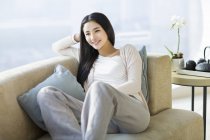 Mujer china sentada en el sofá por ventana en la sala de estar - foto de stock